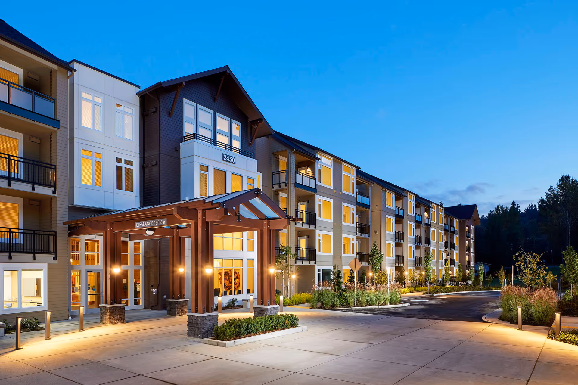 Exterior view of a modern multi-story senior living facility building at dusk with warm interior lights visible through large windows, a covered entrance with wooden beams, landscaped greenery, and a paved driveway.