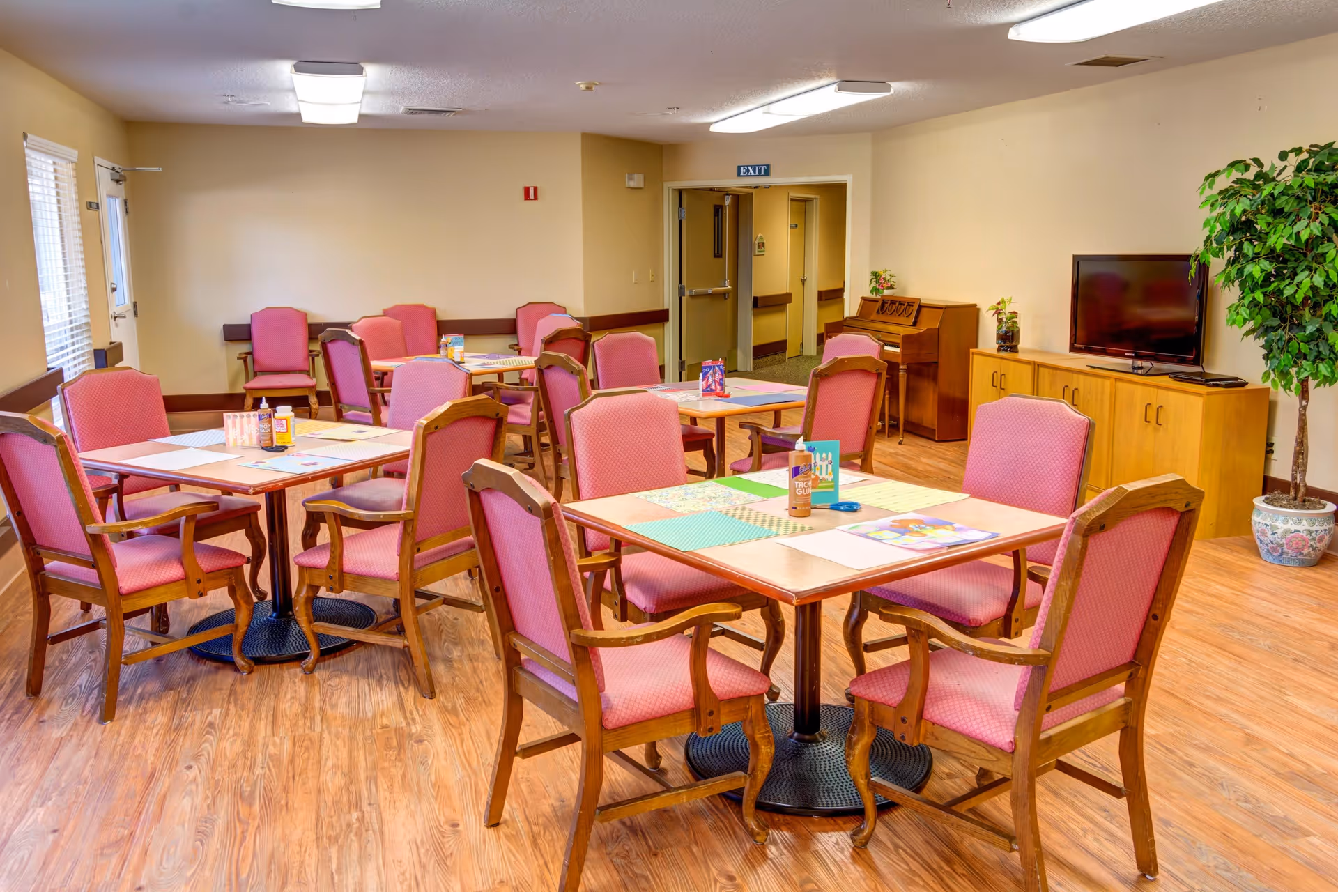 A senior living facility common room with several wooden tables and pink cushioned chairs arranged around them. The room has wood flooring, a television on a wooden cabinet, a piano, a potted plant, and an exit door in the background.