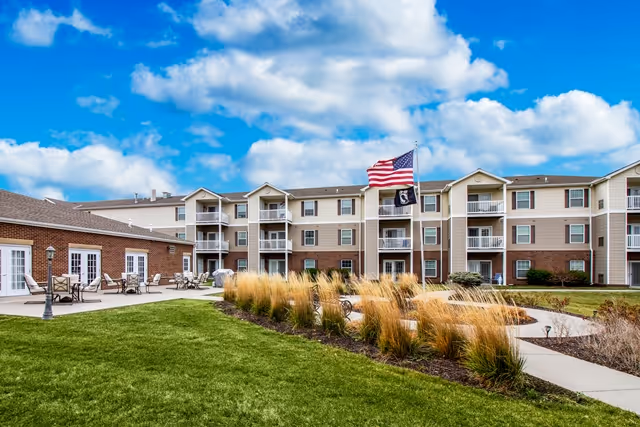 Three-story senior living building with balconies overlooking a landscaped courtyard, patio seating, and flagpoles.