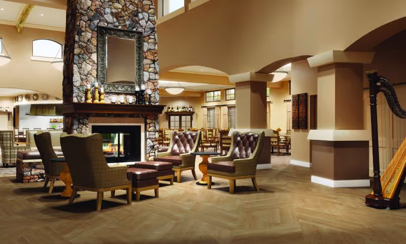 Open communal lounge with a stone fireplace surrounded by armchairs and ottomans, dining tables in the background, and a harp to the side.