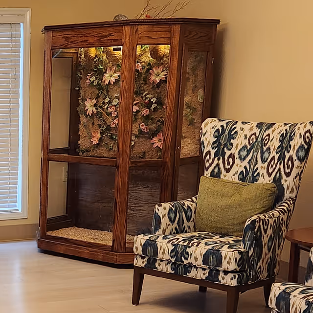 A cozy corner of a room featuring a patterned upholstered armchair with a green cushion, a wooden display cabinet with glass panels containing decorative floral arrangements, and a window with closed blinds.