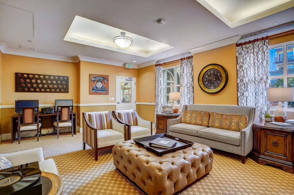A cozy living room area with a beige tufted ottoman in the center, a gray sofa with patterned cushions, two striped armchairs, and two wooden side tables with lamps. The walls are painted yellow with white trim, decorated with a large round clock and artwork. There are two windows with patterned curtains letting in natural light, and a small desk area with two chairs and computers in the corner.