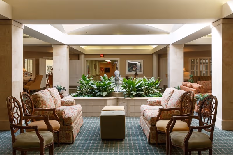 A well-lit indoor seating area with floral upholstered armchairs and wooden chairs arranged around a small ottoman. In the center, there is a planter with green plants and a decorative bird statue. The space has beige columns and a skylight ceiling, with additional seating and framed artwork visible in the background.