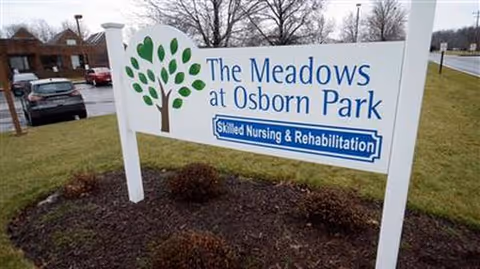 Outdoor sign for The Meadows at Osborn Park with trees, shrubs, and a parking lot nearby.