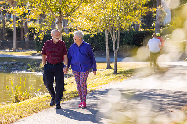 An elderly couple holding hands and walking along a paved path beside a pond in a park-like setting with trees and sunlight filtering through the leaves. In the background, two other people are sitting on a bench.