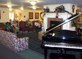 A cozy common area in a senior living facility featuring a black grand piano in the foreground, floral patterned couches, several armchairs, a fireplace decorated with greenery and flowers, and a few elderly residents seated and conversing in the background.