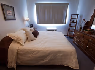 A neatly made bed with white bedding and multiple pillows in a bedroom. There is a wooden dresser with a mirror on the right side, a small wooden ladder shelf with decorative items, a window with closed blinds, and a floor lamp beside the bed. A framed picture hangs on the wall to the left.