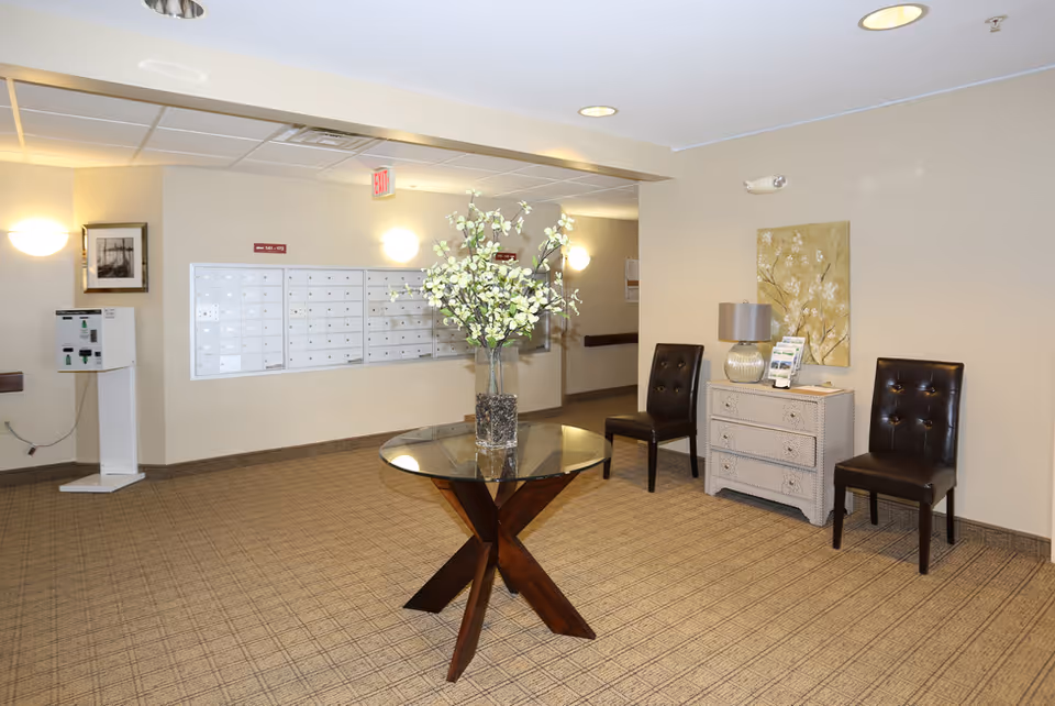 Interior view of a senior apartment facility lobby area with a round glass table holding a vase of white flowers in the center. There are two dark brown leather chairs on either side of a small white dresser with a lamp and brochures on top. A wall-mounted mailbox unit is visible in the background along with soft lighting and beige walls.
