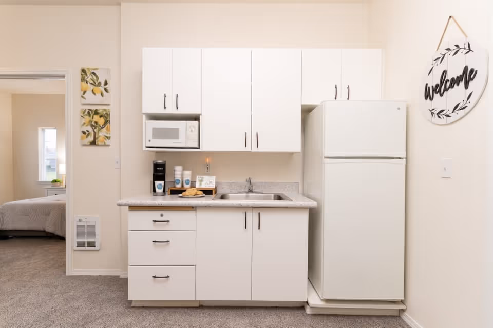 Compact kitchenette with white cabinets, microwave, sink, coffee maker and refrigerator, with a bedroom visible through an open doorway.