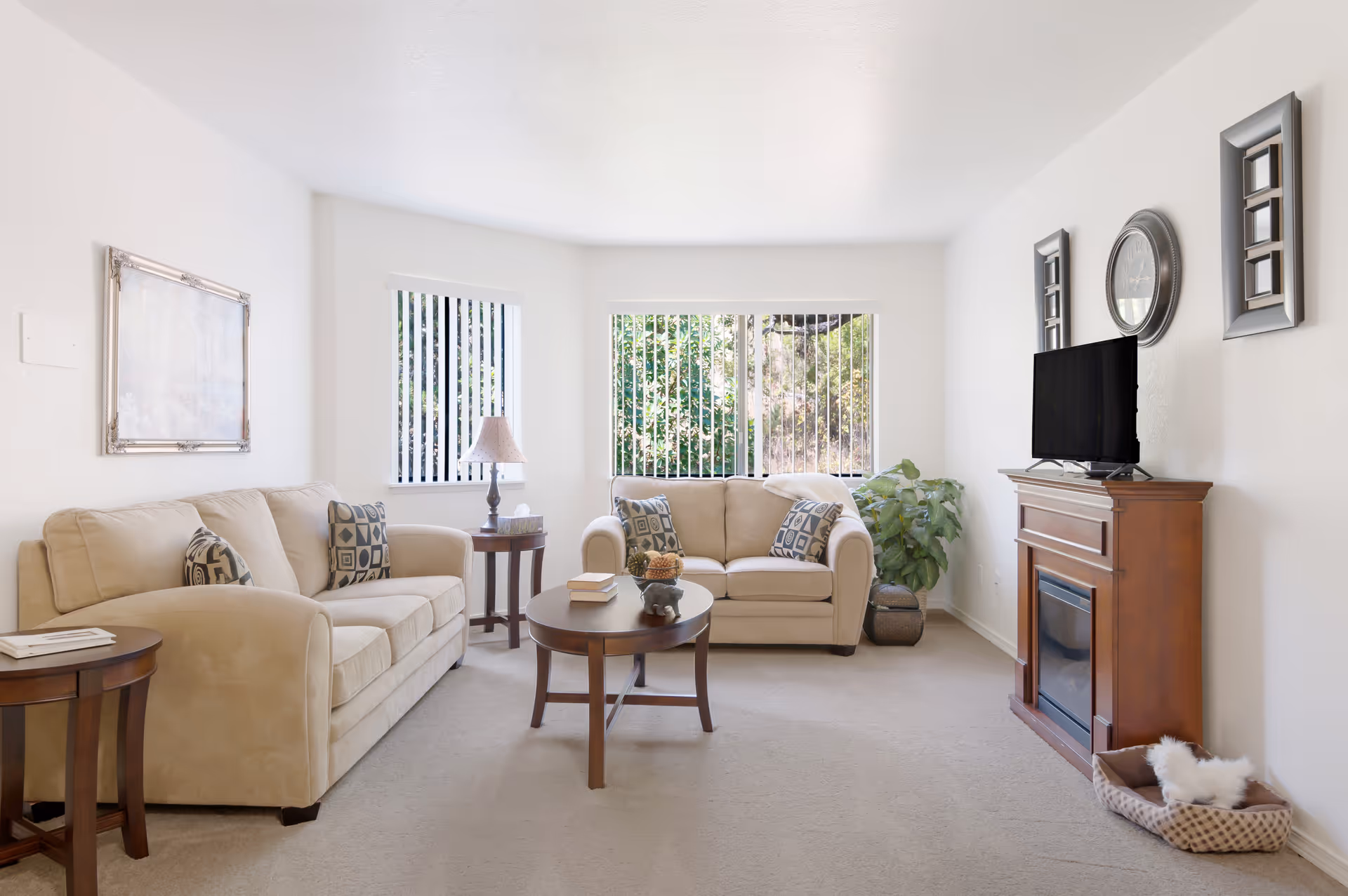 Bright living room with two beige sofas, a round coffee table, a TV on a wooden console with a fireplace, and large windows with vertical blinds.
