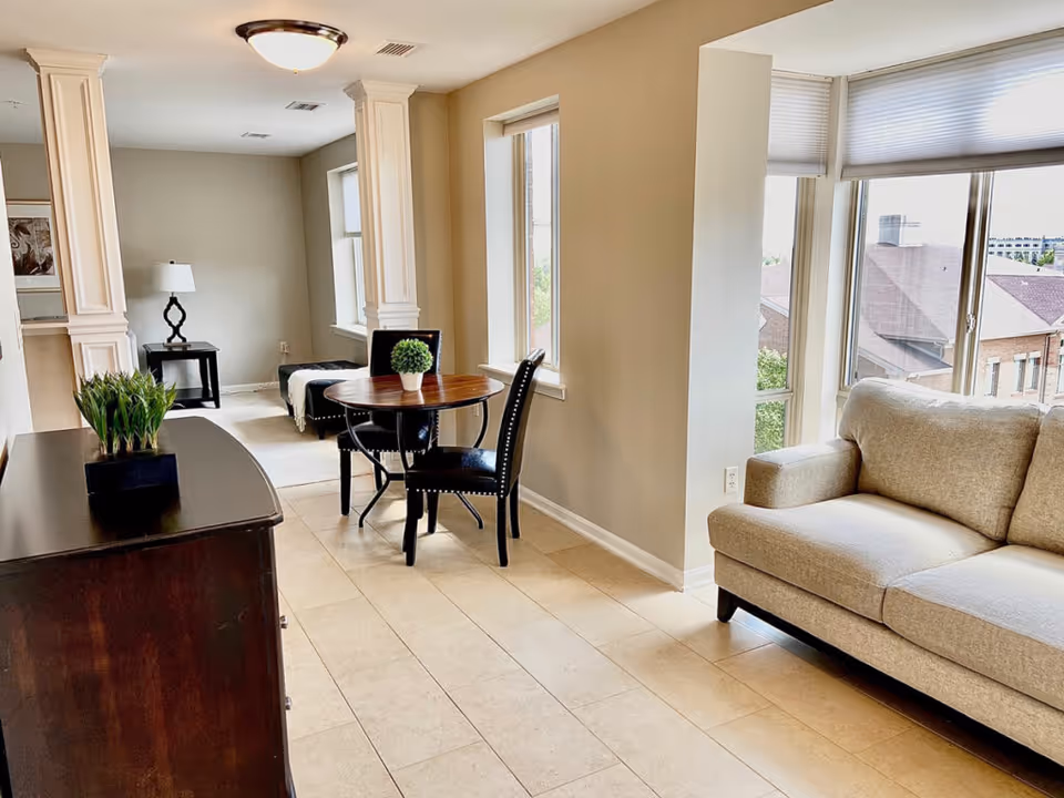A bright and clean interior space featuring a beige sofa near large windows with blinds, a small round wooden table with two black chairs, a dark wooden cabinet with a small green plant, and a side table with a lamp in the background. The room has tiled flooring and light-colored walls.
