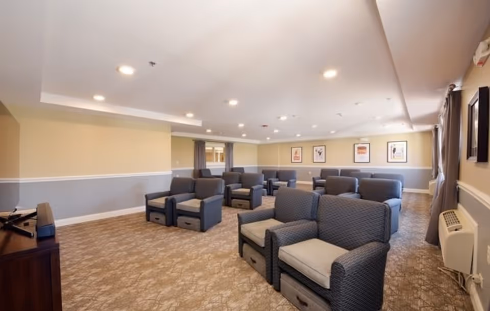 A spacious common area with multiple cushioned armchairs arranged in rows facing a TV on a wooden stand. The room has beige and gray walls, carpeted floor, recessed ceiling lights, and framed artwork on the far wall. Windows with curtains are on the right side.