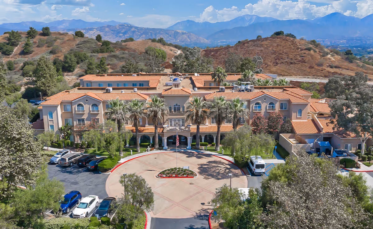 Aerial view of a Mediterranean-style senior living building with a circular front driveway, palm trees, parked cars, and hills in the background.