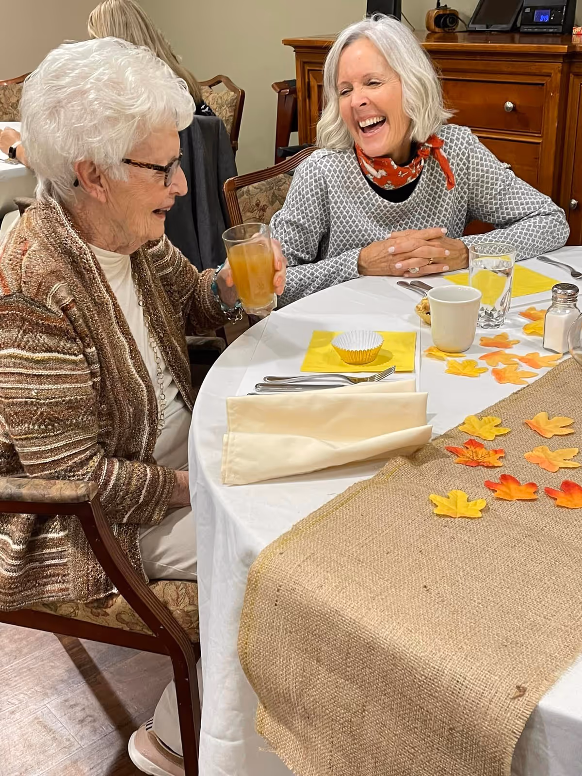 Two elderly women sitting at a dining table decorated with a burlap table runner and artificial autumn leaves. One woman is holding a glass of iced tea and smiling, while the other woman is laughing. The table is set with yellow napkins, utensils, a cup, a glass of water, and a salt shaker. The setting appears to be a cozy indoor dining area.