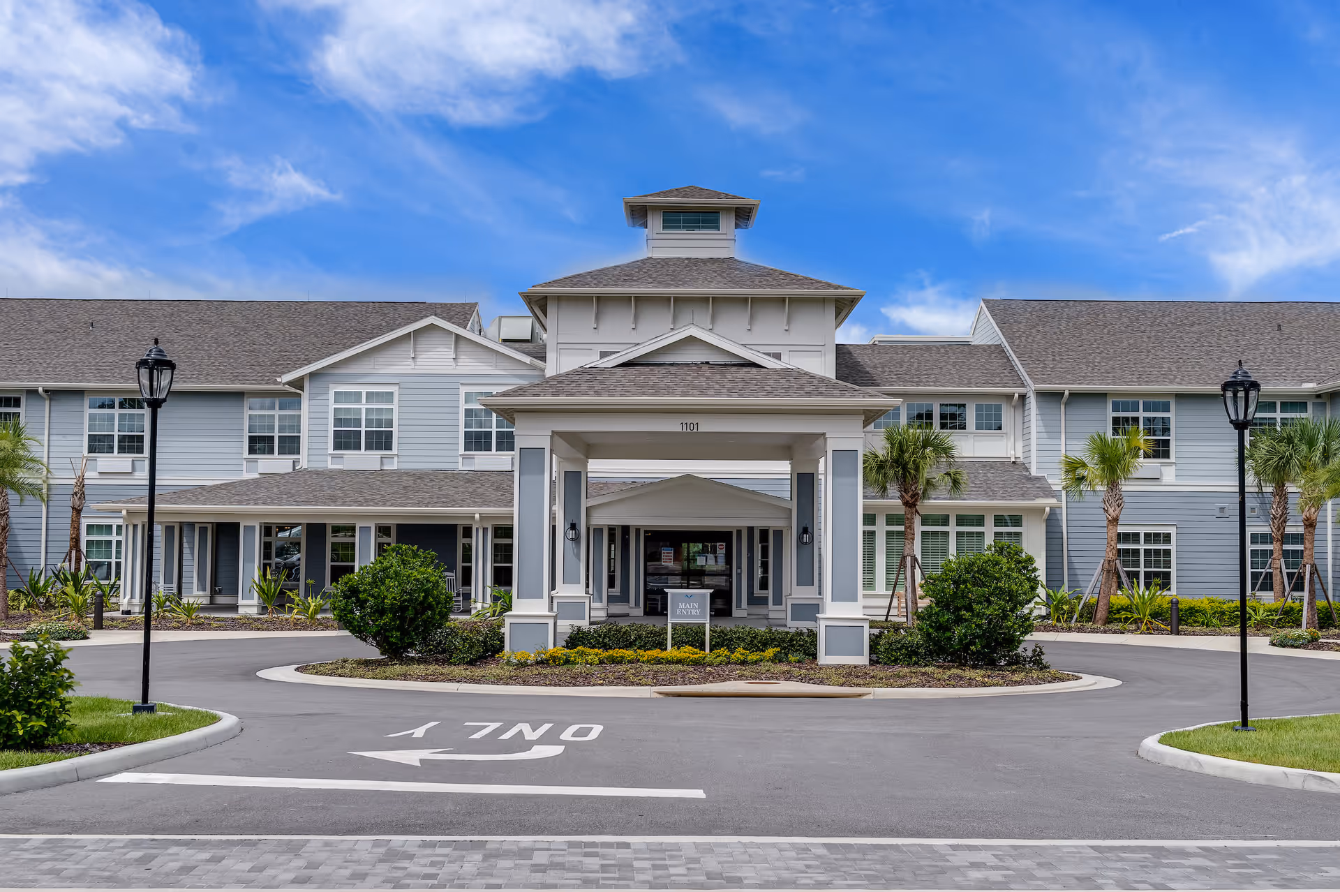Front exterior view of a senior living facility named Sancerre at Orange City, featuring a large covered entrance with the address number 1101 above it, surrounded by landscaped bushes and palm trees, with a clear blue sky in the background.