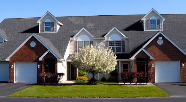 Front exterior view of a residential building with two attached units, each having a garage, dormer windows, and a small porch. A flowering tree and neatly trimmed bushes are in the center lawn area under a clear blue sky.