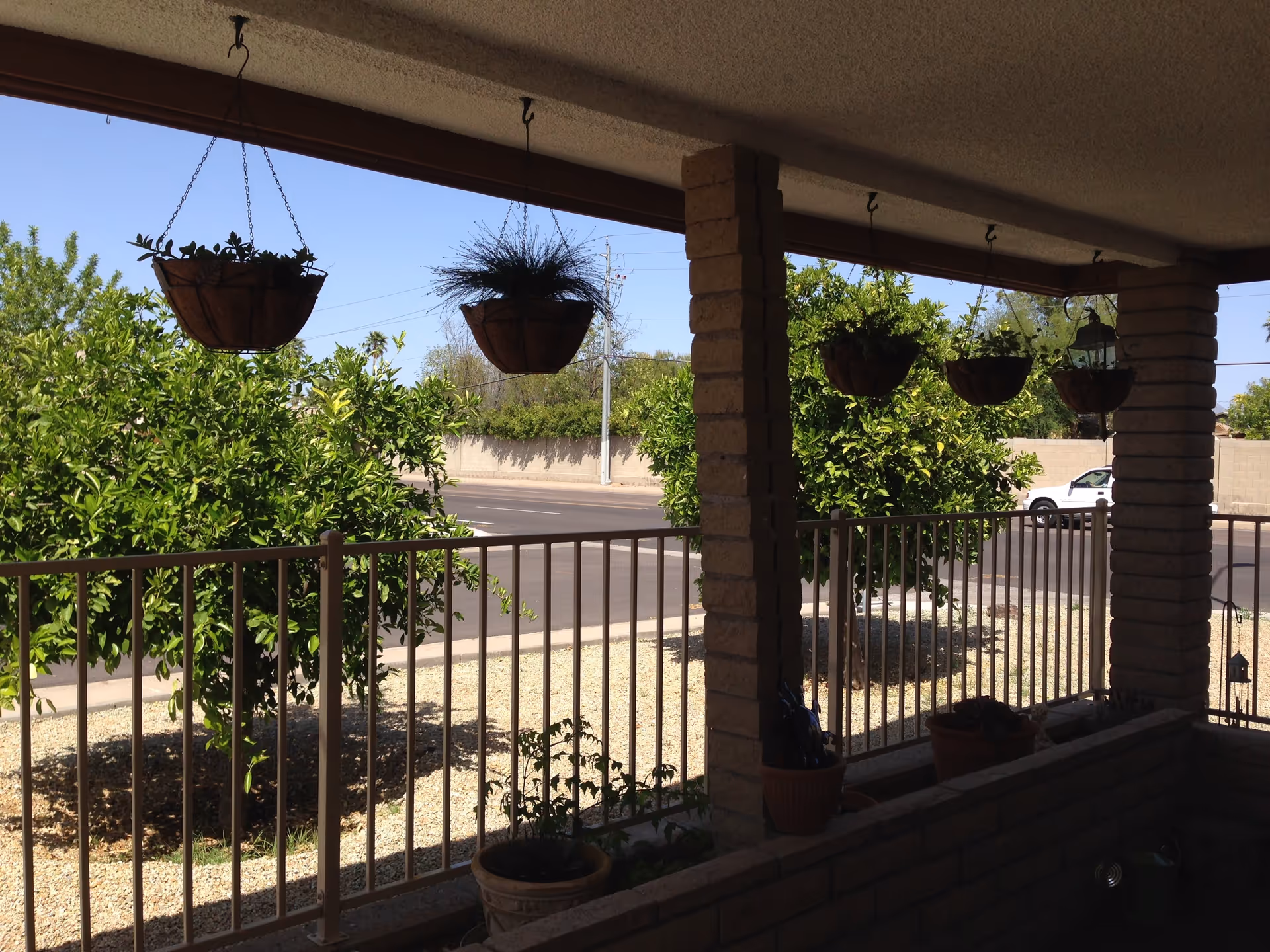 View from a covered front porch with hanging planters and a railing overlooking a street and trees.