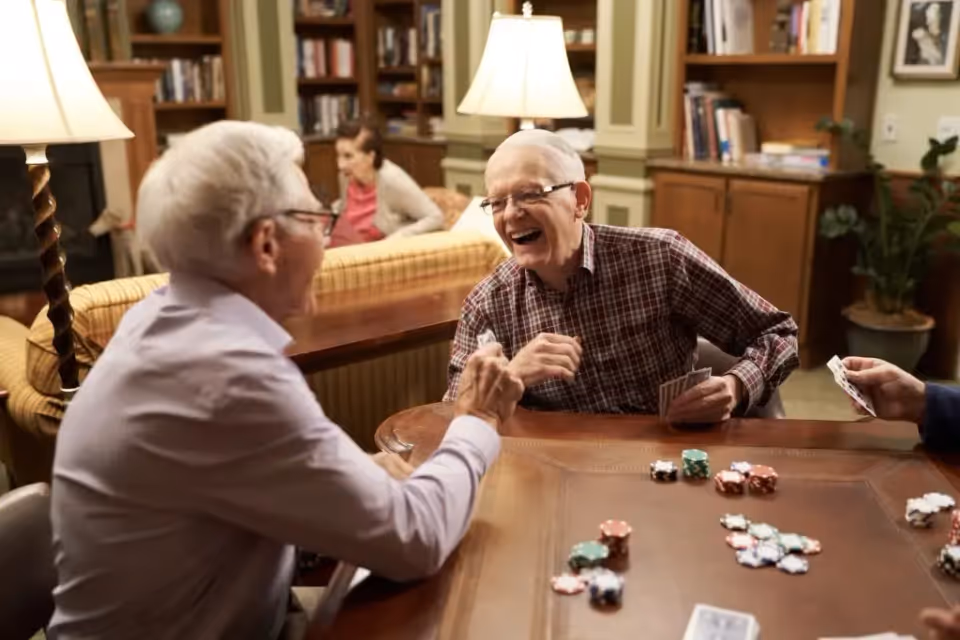 Two elderly men sitting at a wooden table playing cards and poker chips, laughing and enjoying each other's company in a cozy room with bookshelves, lamps, and a woman sitting on a couch in the background.