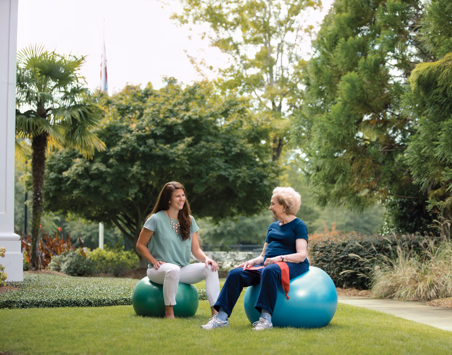 An elderly woman and a younger woman sitting on large exercise balls on a grassy lawn outdoors, surrounded by trees and greenery, engaging in a friendly conversation.