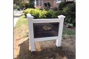A black sign with gold lettering and a gold emblem mounted on white posts in front of a brick building. The sign reads 'Lydia H. Roper Home' and is surrounded by grass and bushes.