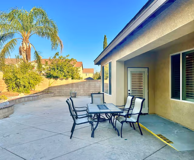 Outdoor patio area with a square table and four cushioned chairs on a concrete surface. The patio is adjacent to a beige building with a door and window. There are palm trees and other greenery along a brick wall in the background under a clear blue sky.