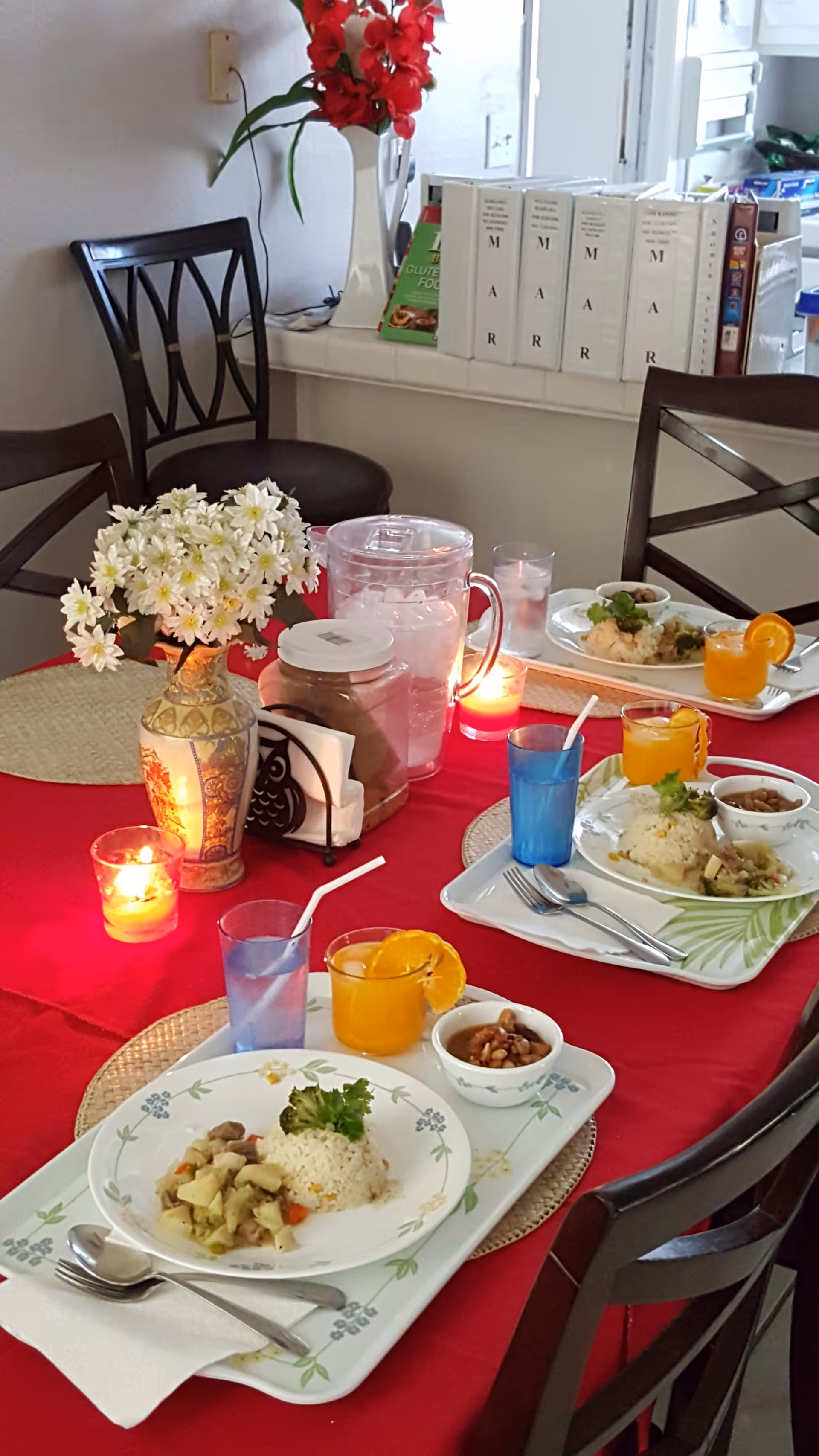 A dining table set with a red tablecloth and three trays of food, each containing a plate of rice with vegetables, a small bowl of beans, a glass of orange juice with an orange slice, and a glass of water with a straw. The table also has a vase with white flowers, a container with napkins, a pitcher of ice water, and two lit candles. In the background, there are chairs and a window sill with books and other items.