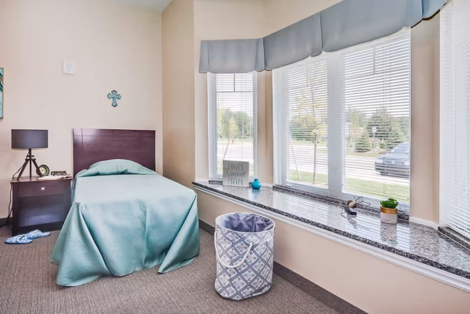 A small, neatly arranged bedroom in an assisted living facility with a single bed covered in a light blue bedspread. Next to the bed is a dark wooden nightstand with a lamp and a small clock. A pair of slippers is on the carpeted floor beside the nightstand. The room has a large bay window with blinds, a granite window sill decorated with a small plant, a blue vase, and a sign that reads 'Mom Sweet Mom'. A patterned laundry basket is placed on the floor near the window.