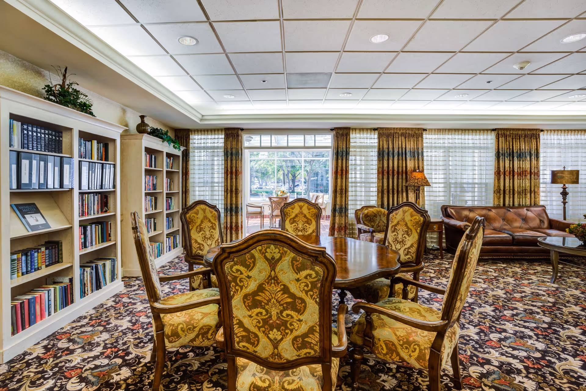 Elegant senior living common room with a wooden table and upholstered chairs, bookshelves, patterned carpet, and seating by large windows.