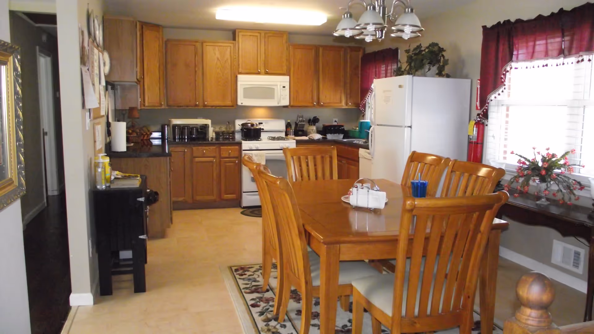 A kitchen and dining area with wooden cabinets, a white refrigerator, a white microwave, and a white stove. A wooden dining table with six matching chairs is placed on a floral rug. The room has beige tiled flooring, two windows with red curtains, and a small table with a flower arrangement near the windows.