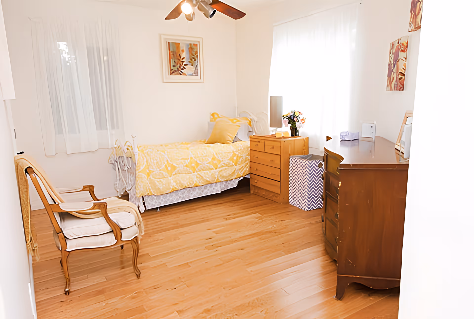 A bright bedroom with wooden flooring, a single bed with yellow and white bedding, a wooden dresser with a lamp and flowers, a wooden chair with cushions, and sheer white curtains on the windows.