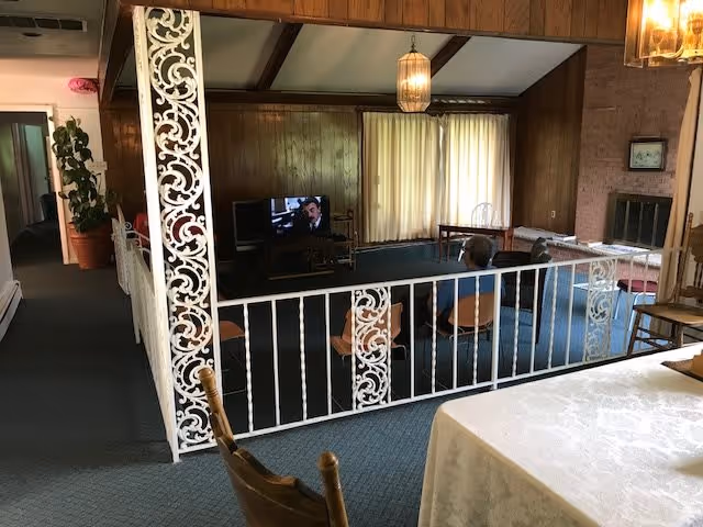 Interior view of a senior living facility common area with a person sitting on a chair watching television. The room has wood-paneled walls, a carpeted floor, a fireplace, and large windows with curtains. There is a decorative white metal railing separating the seating area from the adjacent hallway and dining area.