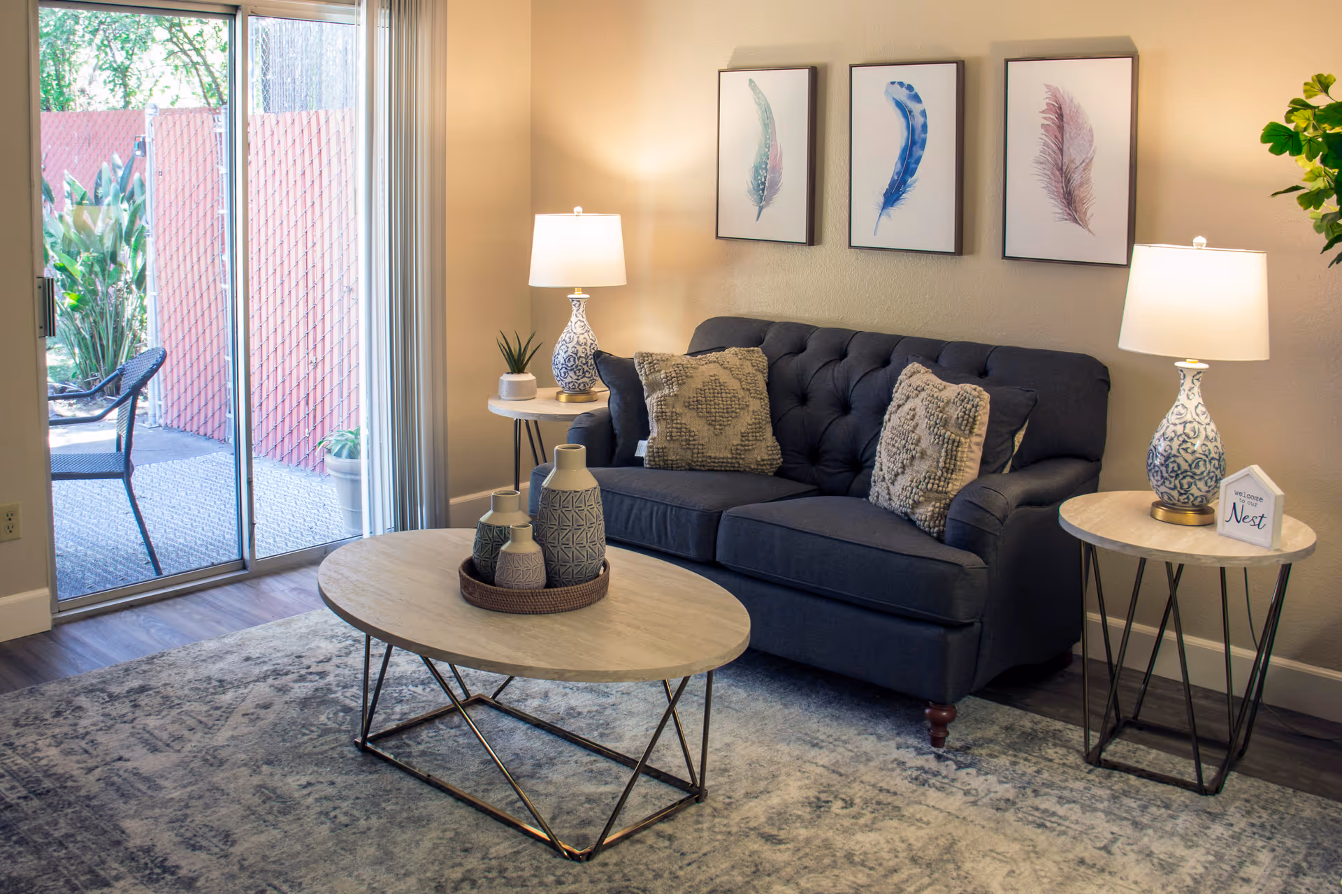 A cozy living room with a dark blue tufted sofa adorned with two textured pillows. There are two side tables, each with a decorative lamp and small decor items. A round coffee table with decorative vases sits on a patterned rug. Three framed feather artworks hang on the wall above the sofa. Sliding glass doors open to an outdoor patio with a chair and plants.
