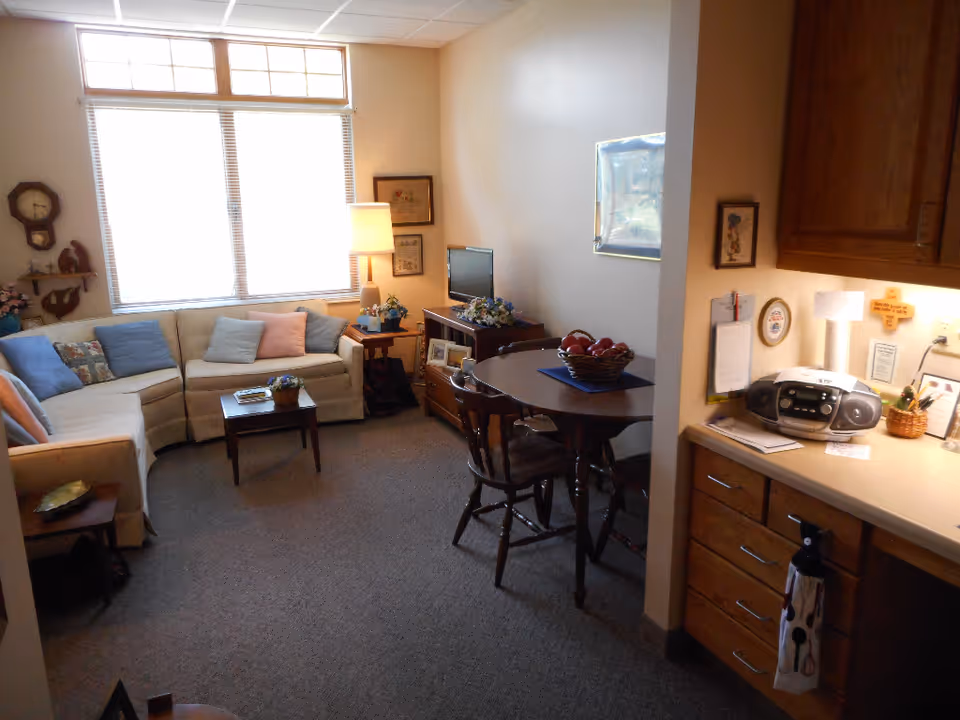 A cozy living room area with a beige sectional sofa adorned with various cushions, a small wooden coffee table with a plant on it, a side table with a lamp and flowers, a small TV on a wooden stand, and a round dining table with chairs and a bowl of apples. The room has a large window with blinds, carpeted floor, and wooden cabinets with a countertop holding a radio and some papers.