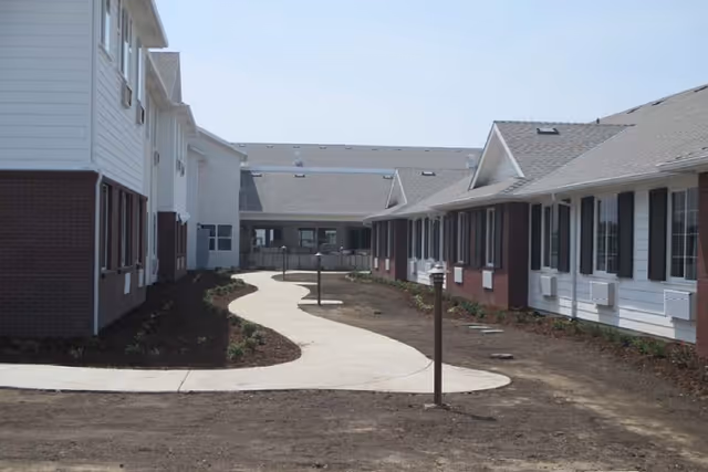 A winding concrete pathway between two single-story buildings with white siding and brick accents. The buildings have multiple windows with shutters and air conditioning units beneath them. The ground around the pathway appears to be bare soil with some small plants near the buildings. The sky is clear and blue.
