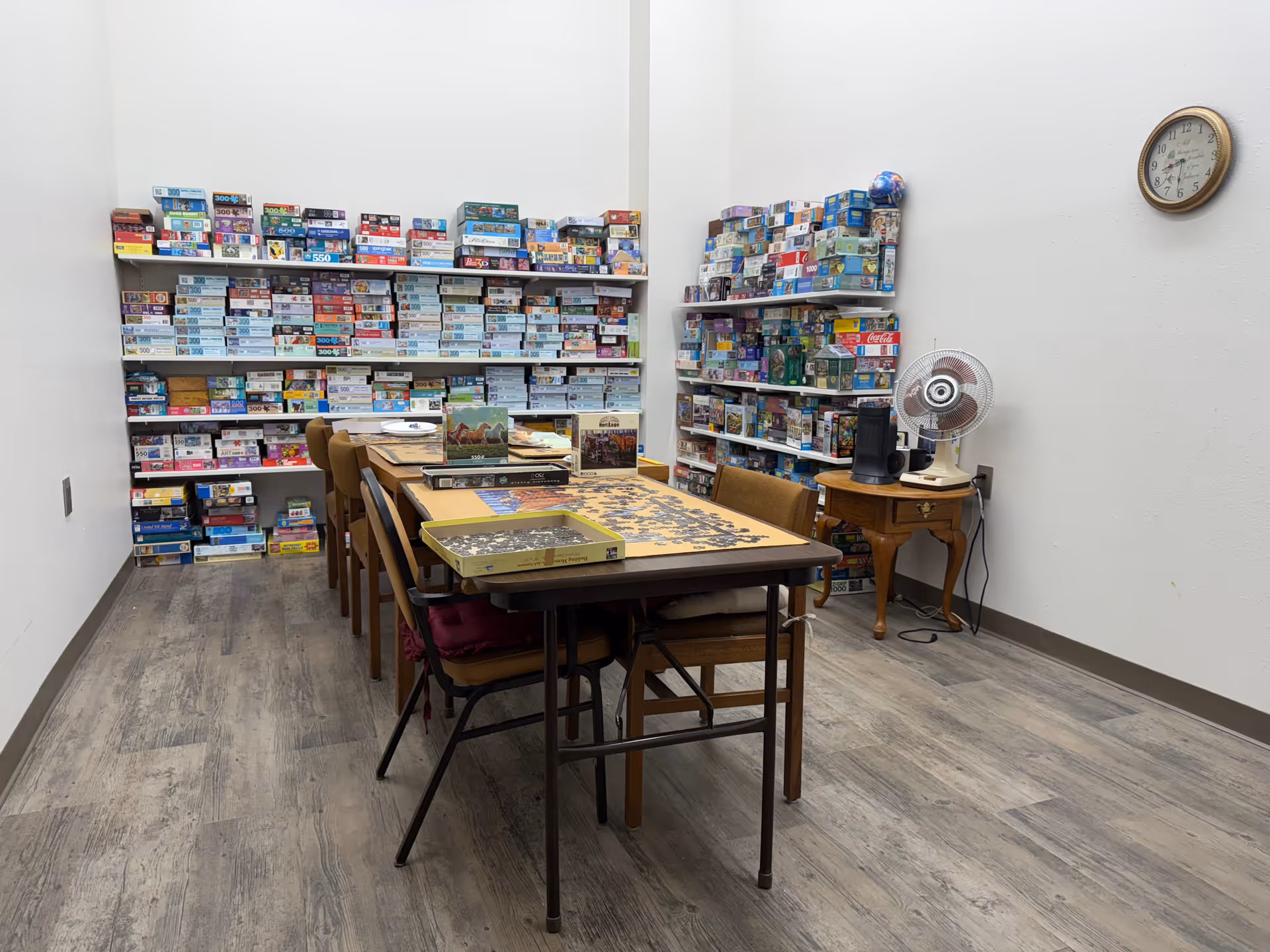A room with shelves filled with numerous puzzle boxes along the back and right walls. In the center, there is a long table with chairs around it, and a partially completed puzzle on the table. A small wooden side table holds a fan and a speaker, and a clock is mounted on the wall above it.