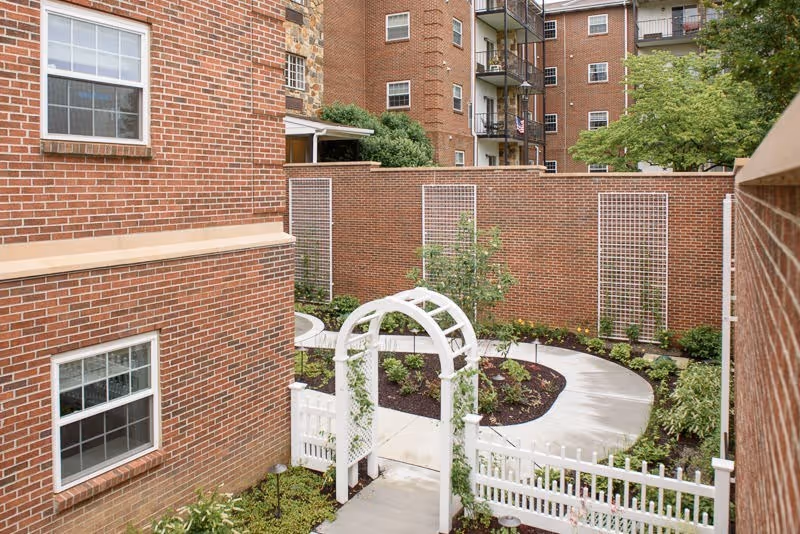 A small enclosed garden area surrounded by brick buildings with windows. The garden features a white wooden archway with climbing plants, a white picket fence, a curved concrete pathway, and various green shrubs and plants along the edges. In the background, there are taller brick apartment buildings with balconies and trees.