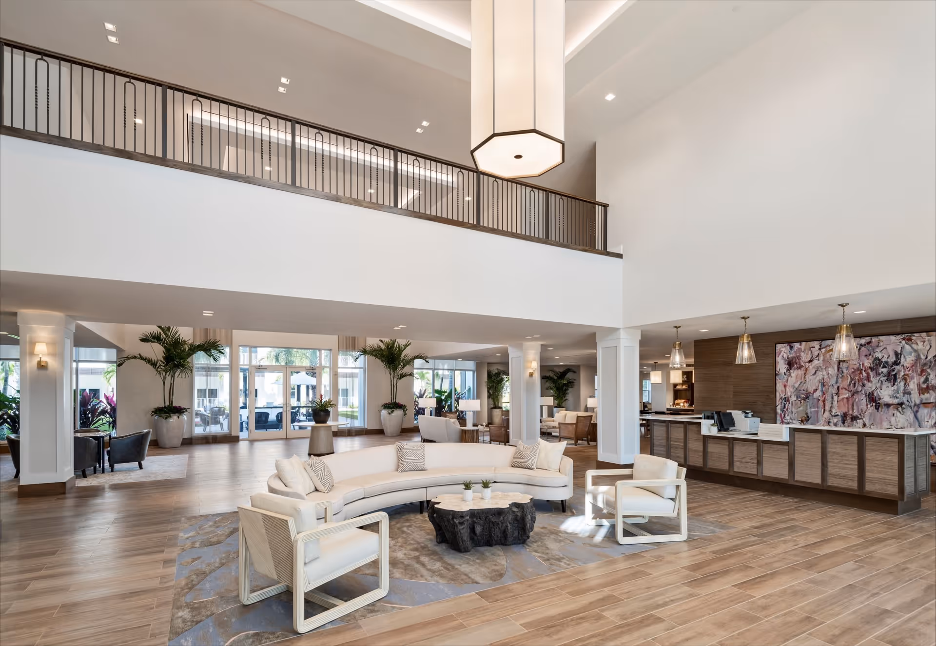 Spacious and modern senior living facility lobby with a high ceiling, a large curved white sofa, two armchairs, a unique wooden coffee table, and a reception desk with pendant lights and abstract artwork behind it. Large windows and glass doors allow natural light to fill the space, with potted plants adding greenery throughout.