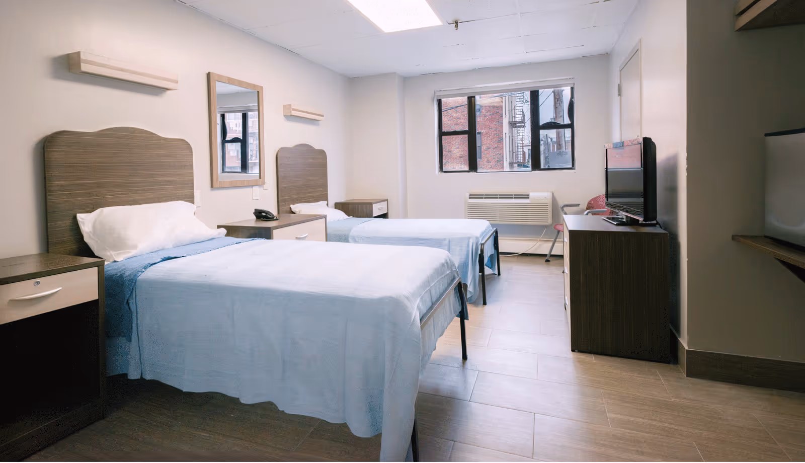 A simple, clean bedroom with two single beds covered in light blue bedspreads. Each bed has a wooden headboard and a white pillow. Between the beds is a small nightstand with a telephone. A rectangular mirror is mounted on the wall above the nightstand. There is a window on the far wall with a view of a brick building outside. Below the window is an air conditioning unit. On the right side of the room, there is a wooden dresser with a flat-screen TV on top and a pink chair next to it. The floor is covered with light brown tiles, and the walls are painted white.