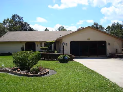 Single-story assisted living facility front yard with manicured lawn, shrubs, and a driveway.