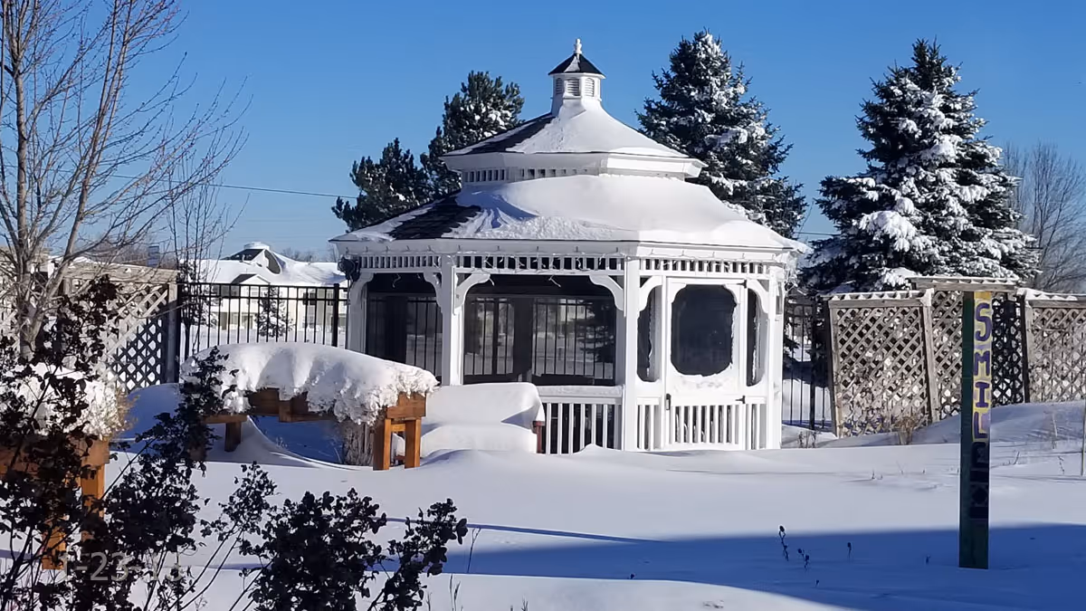 A white gazebo covered in snow in a snow-covered yard with evergreen trees and a colorful 'SMILE' post.