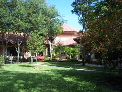 Exterior view of a pinkish-beige building with a red-tiled roof, surrounded by trees, a lawn, and benches along a walkway.