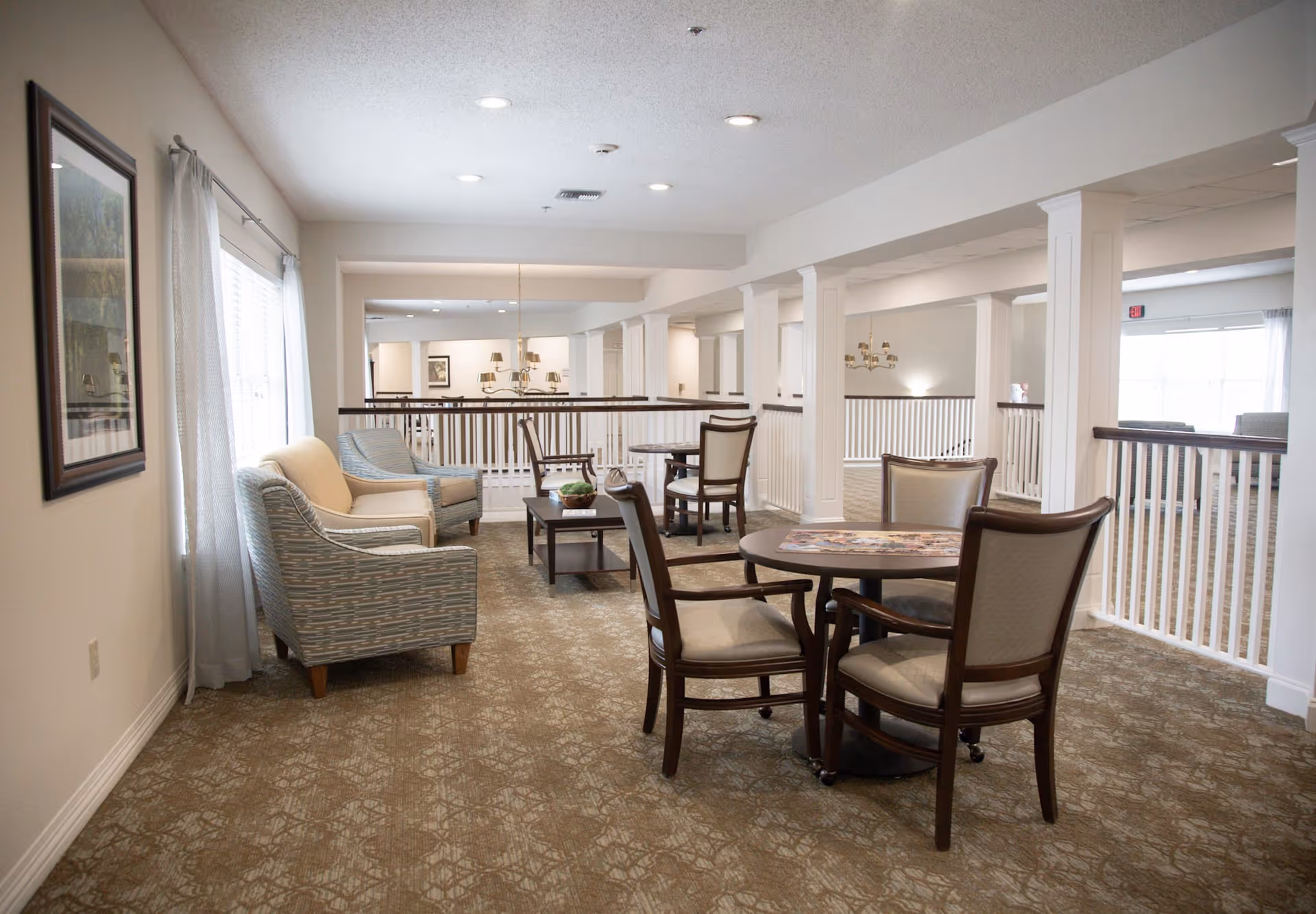 A bright and spacious common area in a senior living facility featuring comfortable armchairs and sofas arranged around coffee tables, with additional round tables and chairs for seating. The space has carpeted floors, white walls, large windows with sheer curtains allowing natural light, and white railings with columns separating the area from a lower level.