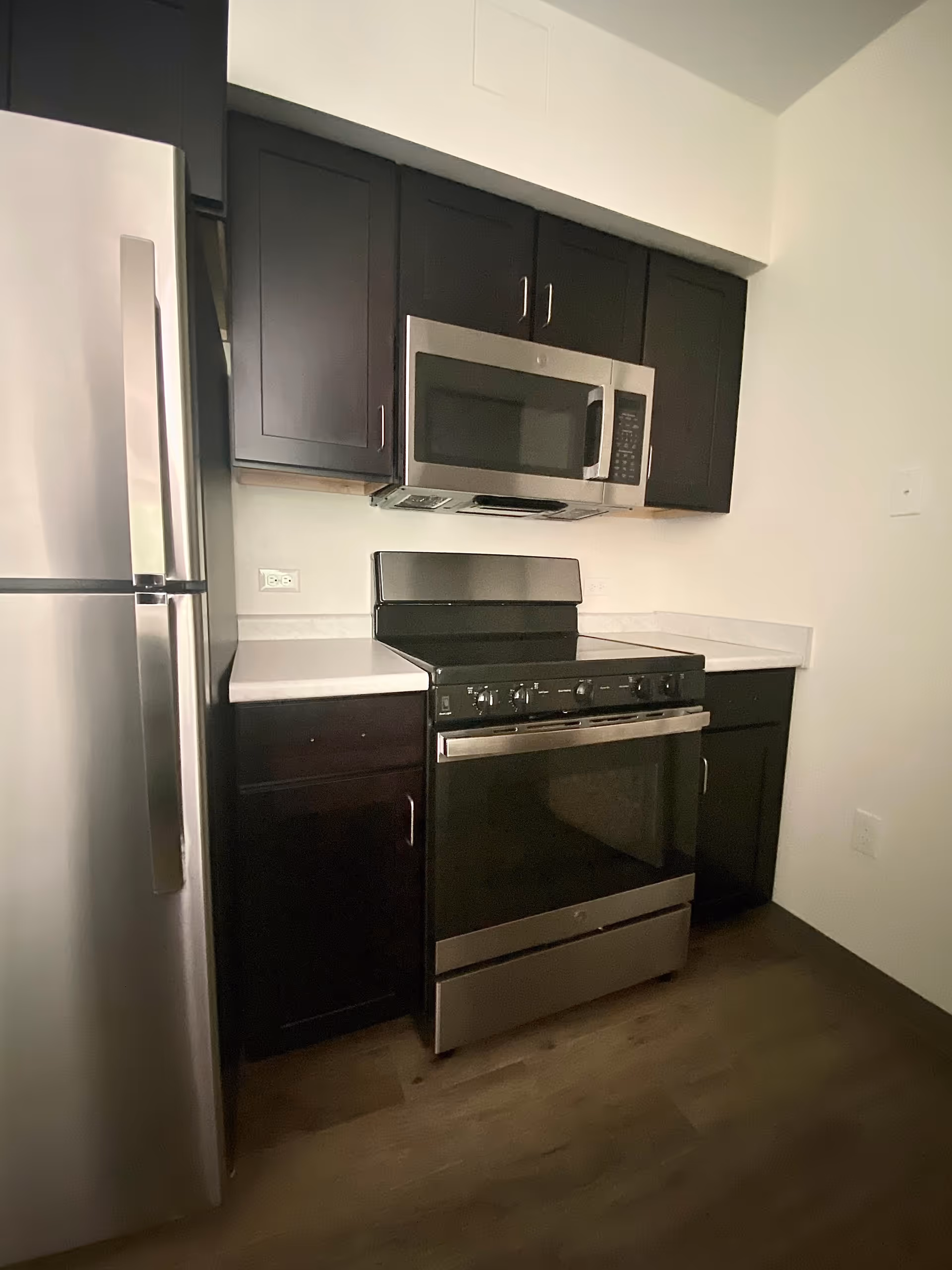 A modern kitchen corner featuring stainless steel appliances including a refrigerator, microwave, and stove with dark wood cabinets above and below white countertops, and wood flooring.