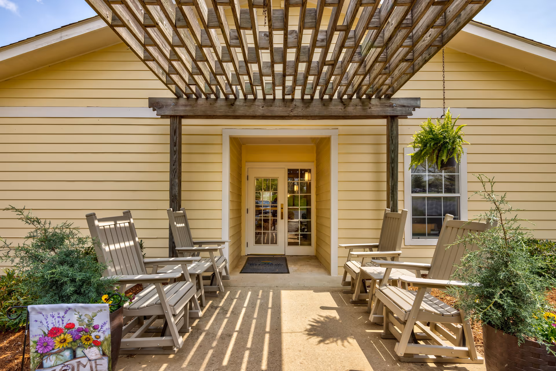 Outdoor seating area with beige wooden rocking chairs arranged on both sides of a concrete pathway leading to a glass double door entrance of a yellow building. A wooden pergola overhead casts striped shadows on the ground. There are green plants and a hanging fern near the entrance.