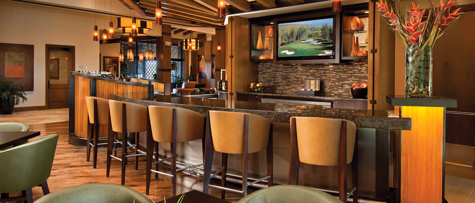 Interior view of a modern lounge area with a curved bar counter and six tan leather bar stools. The space features warm wooden flooring, pendant lighting, and a flat-screen TV mounted on a wall with decorative shelving and a mosaic tile backsplash. Green upholstered chairs and tables are visible in the foreground, along with a tall vase of flowers on a lit pedestal.