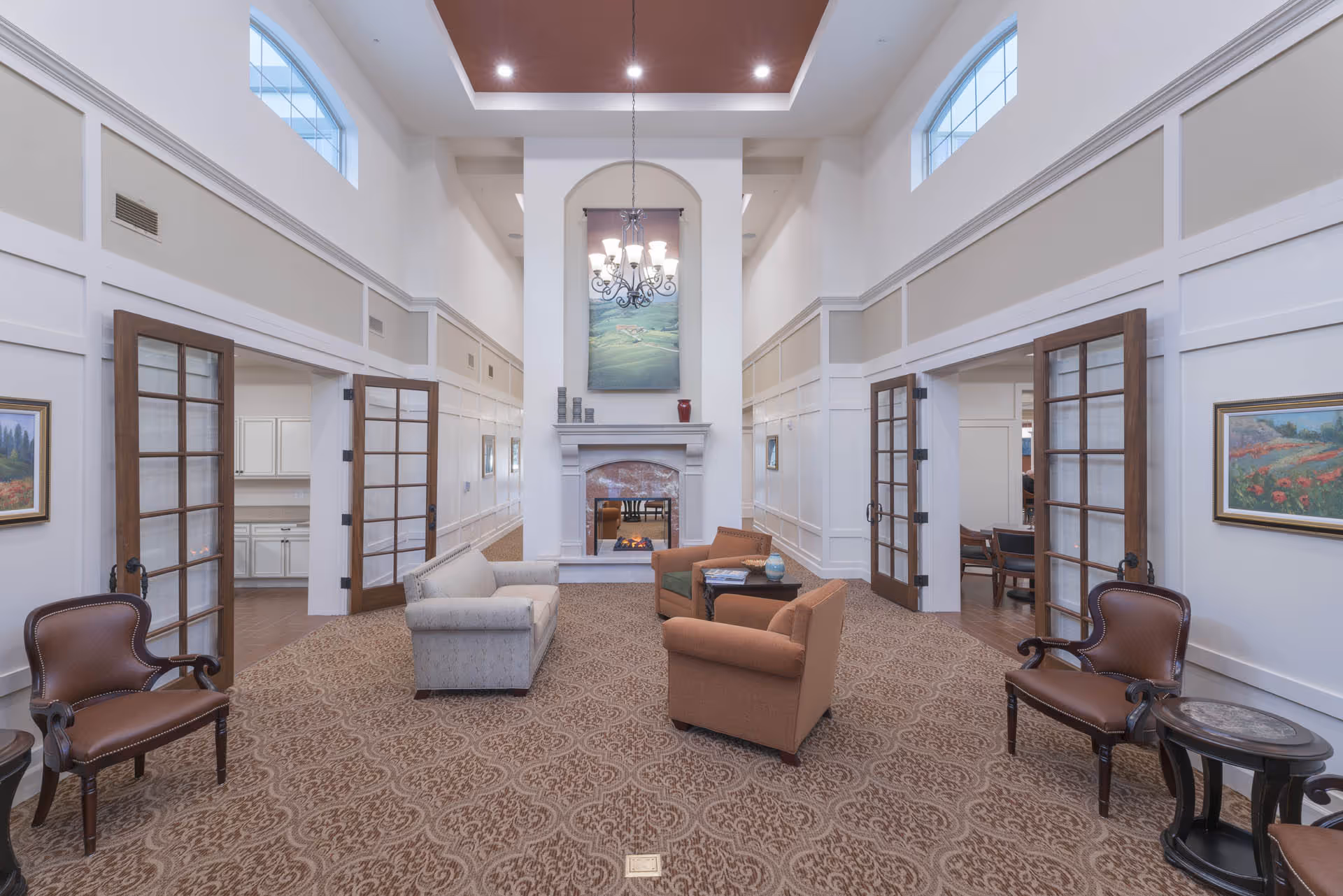 A spacious, well-lit living room with high ceilings and large windows near the ceiling. The room features a central fireplace with a painting above it, surrounded by a beige and brown patterned carpet. There are two brown armchairs and a beige loveseat arranged around a small table in front of the fireplace. On either side of the room, there are wooden framed glass doors leading to other rooms, and the walls are decorated with framed paintings.