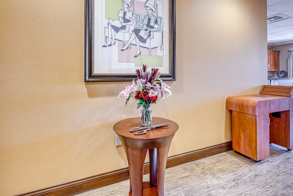 A small wooden round table with a glass vase holding a bouquet of pink and red flowers placed against a beige wall. Above the table hangs a framed artwork depicting two women sitting and reading. To the right, there is a wooden piece of furniture with a curved top, and part of a kitchen area is visible in the background.
