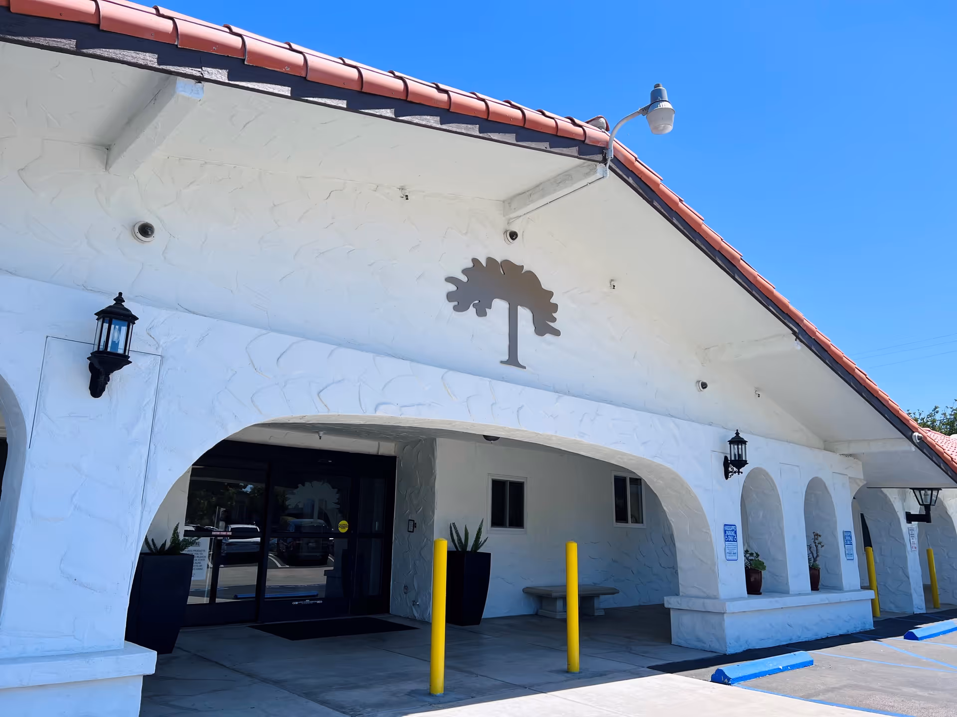 Exterior front entrance of a building with white stucco walls and a red tile roof. The entrance has large arches, black lantern-style wall lights, and a metal tree decoration above the doorway. There are yellow bollards and blue parking curbs in front of the entrance under a clear blue sky.