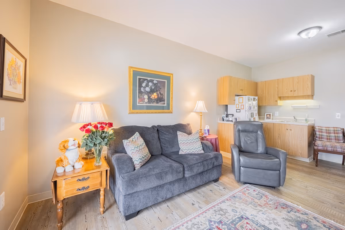 A cozy living room area in a senior living facility featuring a gray sofa with patterned pillows, a wooden side table with a lamp, a vase of red roses, and a stuffed animal. Next to the sofa is a gray recliner chair. The background shows a small kitchenette with wooden cabinets, a refrigerator, and a plaid upholstered chair. The room has light-colored walls and wood flooring with a patterned area rug.