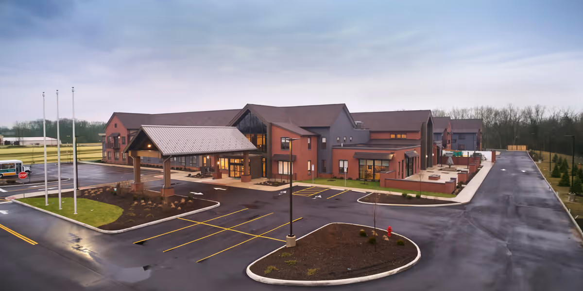 Exterior view of The Patterson senior living facility showing a large two-story building with a covered entrance, multiple windows, and a spacious parking lot with marked parking spaces. The surrounding area includes landscaped sections with grass and small trees under a cloudy sky.
